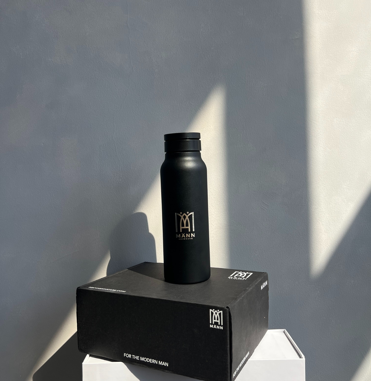 Black water bottle with brand logo on a black box against a gray wall